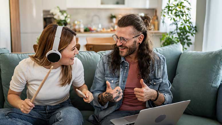 Zwei Personen am gemeinsam Jamen mit Laptop auf dem Schoss, Kopfhörer und Drum-Sticks.