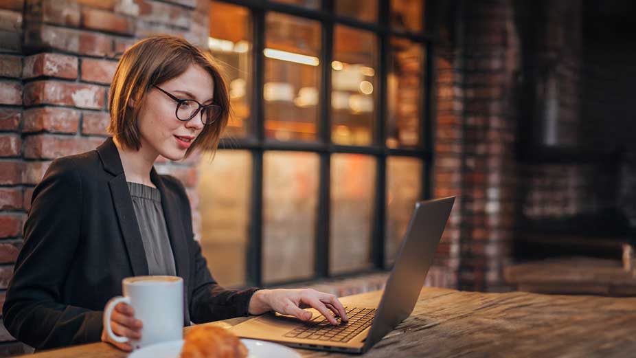 Frau sitzt mit Kaffeetasse in einem trendy Café vor einem Laptop.