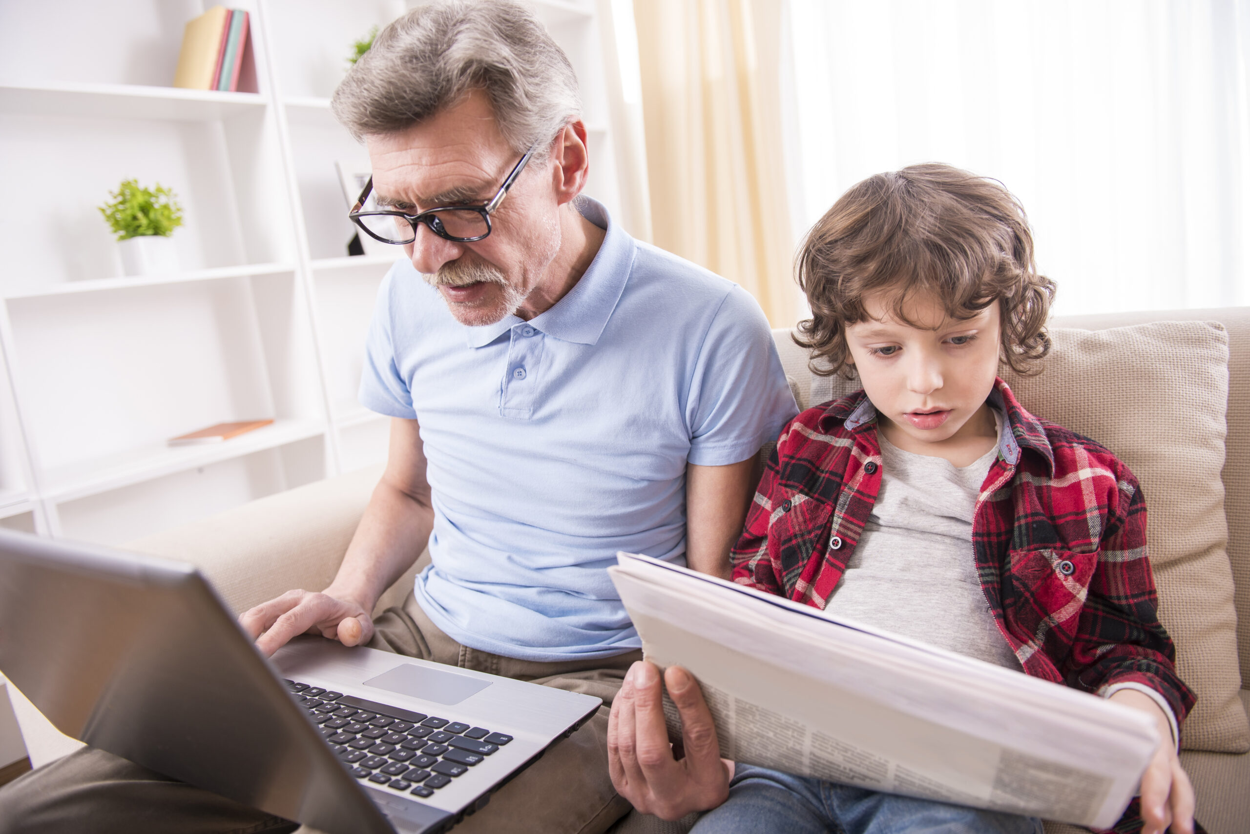 Grand-père et petit-fils sont assis sur le canapé. Le grand-père avec l'ordinateur portable sur les genoux, tandis que le petit-fils lit le journal. 